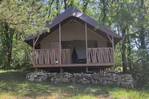 Holz-Campinghütte mit Balkon und Terrasse auf einem Steinsockel im Grünen, umgeben von Bäumen im Wald.