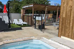 Terrasse am Campingplatz mit Whirlpool, Kiesweg, Rasenfläche und weißen Liegestühlen hinter einem Metallzaun.