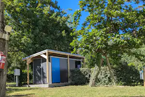 Holzüberdachtes Sanitärgebäude mit blauen Türen auf einem Campingplatz, umgeben von Bäumen und Hecken.