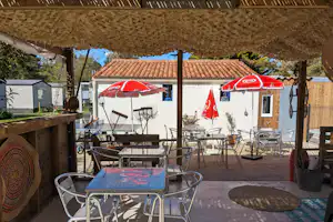 Camping-Terrasse mit Esstisch, Stühlen und zwei roten Sonnenschirmen unter einer Holzüberdachung vor einem weißen Gebäude.