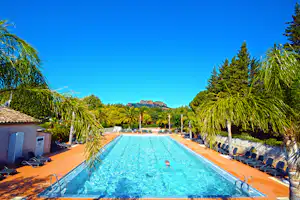 Schwimmbad mit blauem Wasser, Liegestühlen und Palmen auf einer Terrasse, dahinter Bäume und Berge in der Ferne.