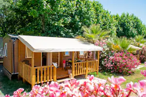 Holzterrasse am Bungalow mit Markise, Sitzgruppe und Blumenbeeten im grünen Garten, neben einem Wohnwagen.