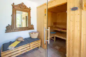 Holzsauna mit Liegebank und offenem Eingang, daneben eine Kommode und ein Spiegel im hellen Raum.