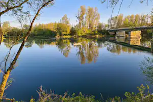 Ruhiger See mit Ruderboot, Bäumen und Uferweg; im Hintergrund eine Brücke über dem Wasser.