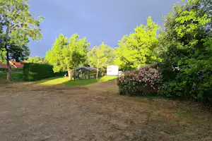 Campingplatz mit Rasenfläche, Hecken und Wohnwagen im Hintergrund, umgeben von Bäumen und einem kleinen Weg.