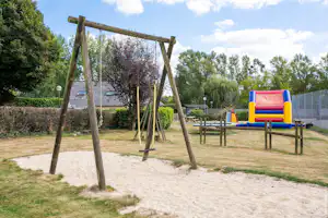 Camping Les Peupliers - Spielplatz auf dem Campingplatz mit Schaukeln, Balancierstange, Tischtennisplatten und Hüpfburg im Hintergrund