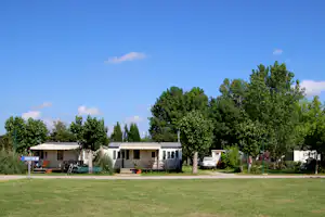 Campingplatz mit Rasenfläche, mehreren Mobilheimen und Wohnwagen sowie Bäumen im Hintergrund.