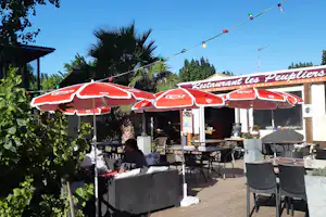 Terrasse eines Restaurants mit roten Sonnenschirmen, Tischen und Stühlen, Holzdeck und Pflanzen vor dem Gebäude.