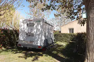 Wohnmobil auf einem Campingplatz-Stellplatz im Grünen, mit Rasenfläche, Hecke und Bäumen neben einem Gebäude.