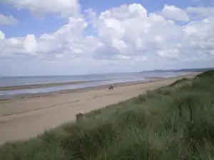 Weite Sandbucht mit Dünen und Meer, Blick vom grünen Hang unter wolkigem Himmel.