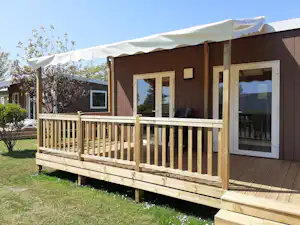 Bungalow mit Holzterrasse und Geländer, Markise am Eingang, große Fensterfront, Rasen und Bäume im Hintergrund.