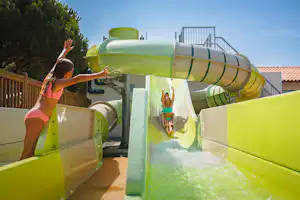 Grüne Wasserrutsche im Campingbereich, Kinder rutschen ins Wasser, Holzgeländer und Bäume im Hintergrund.
