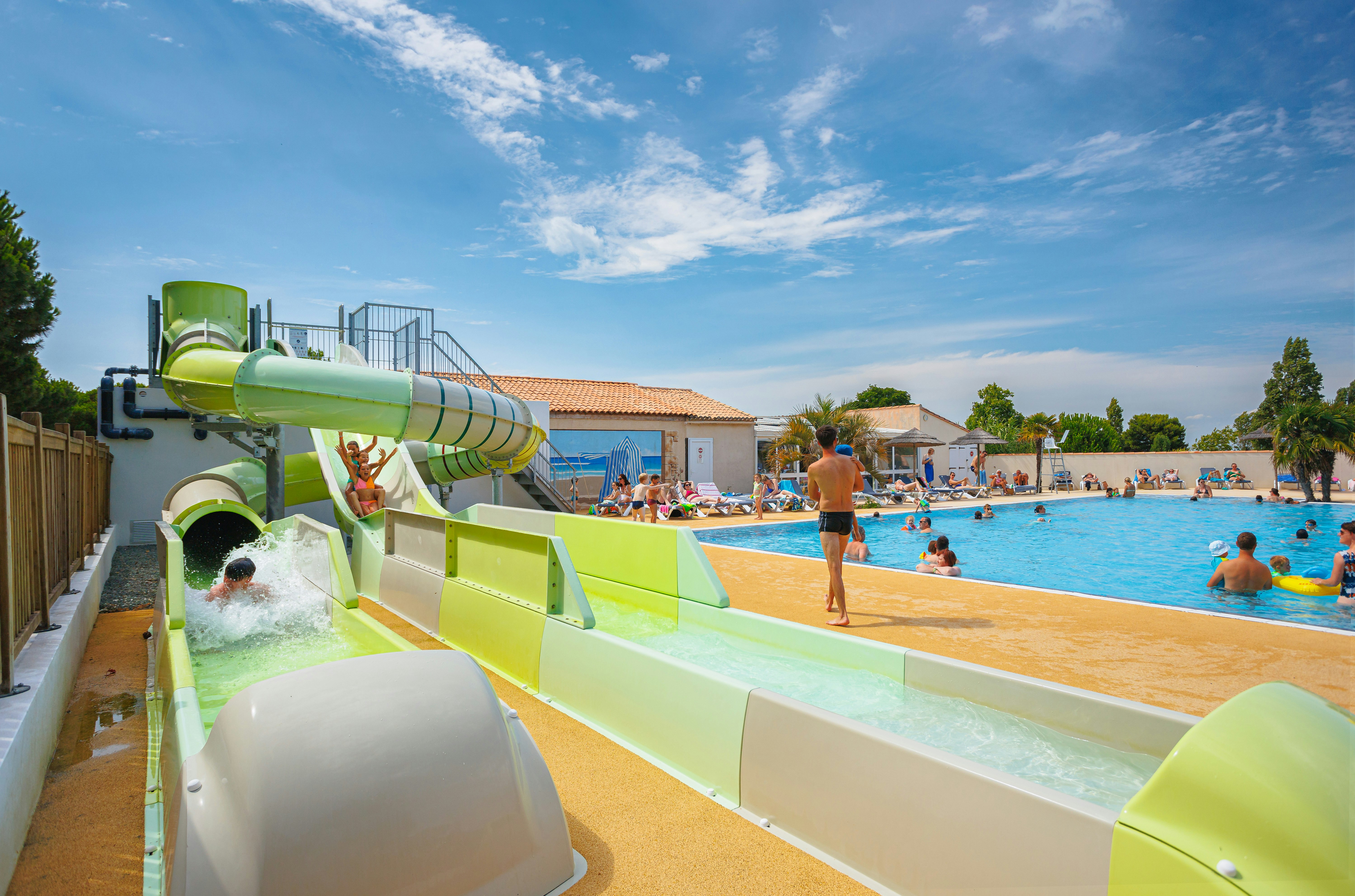 Grüße Wasserrutsche am Camping-Pool, Kinder und Erwachsene im Becken, Liegen und Gebäude im Hintergrund.