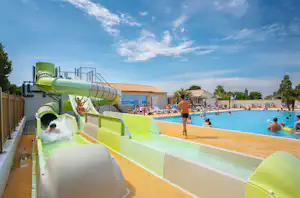 Grüße Wasserrutsche am Camping-Pool, Kinder und Erwachsene im Becken, Liegen und Gebäude im Hintergrund.