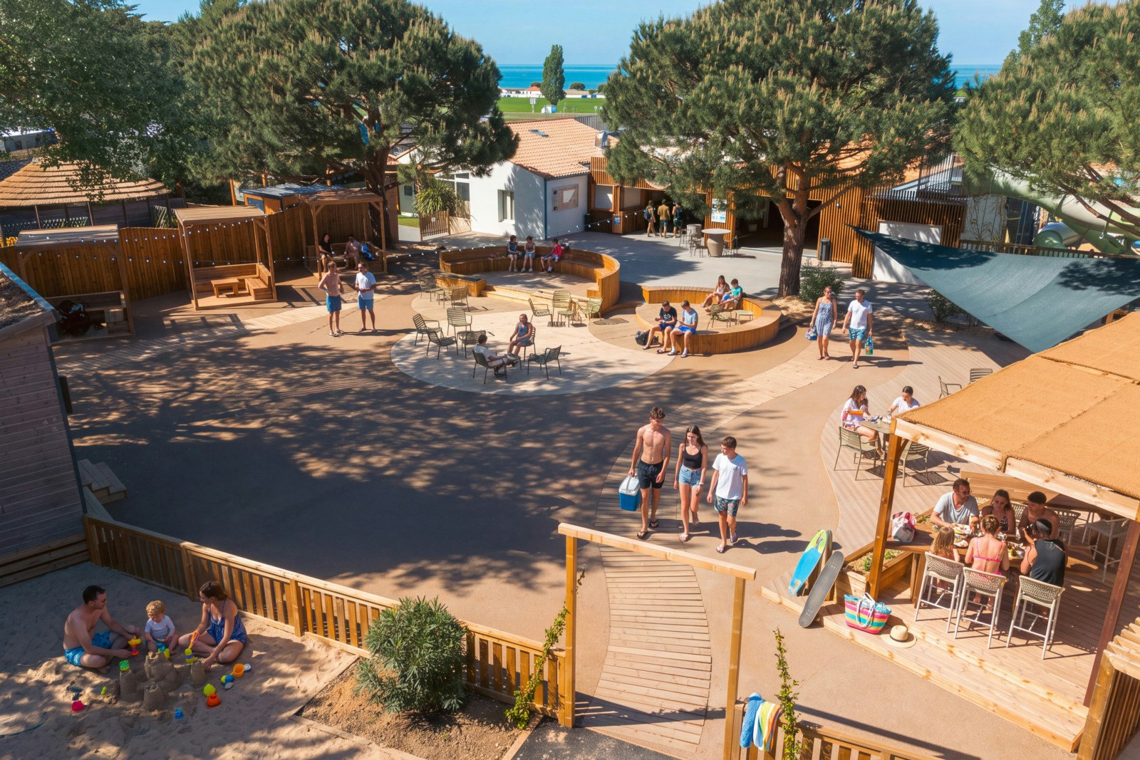 Großer Poolbereich mit Sonnenliegen, Menschen und einem Holzdeck, umgeben von Bäumen und Ferienhäusern im Campingplatz.