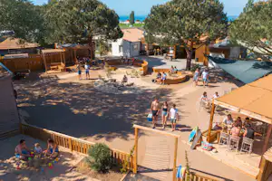 Großer Poolbereich mit Sonnenliegen, Menschen und einem Holzdeck, umgeben von Bäumen und Ferienhäusern im Campingplatz.