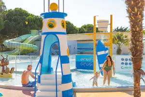 Blauer Spielturm mit Rutsche am Pool, daneben Kinder und Erwachsene im Schwimmbereich, Palmen und Liegen am Rand.