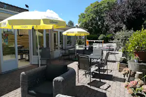 Terrasse am Campingplatz mit Tischen und Stühlen, gelben Sonnenschirmen und Blick auf das Gebäude mit Glastür.
