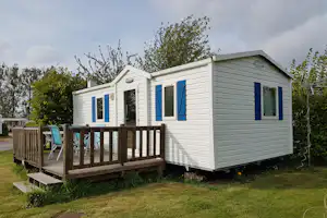 Weißes Mobilheim mit Holzveranda, blauen Fensterläden und Treppe auf einem Campingplatz, umgeben von Rasen und Bäumen.