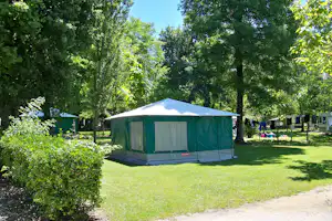 Grünes Zelt auf einer Wiese im Campingplatz, umgeben von Bäumen und Hecken, mit Weg und Sitzbereich im Hintergrund.