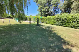 Grüne Campingwiese mit Zaun, Bäumen und Hecke am Rand, im Hintergrund ein Weg und ein Spielbereich.