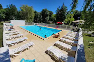 Camping-Pool mit blauem Wasser, mehreren Liegen auf der Terrasse und Personen im Becken, umgeben von Bäumen und Rasen.