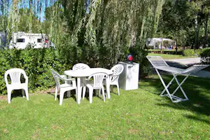 Camping-Garten mit weißen Kunststoffstühlen und Tisch, daneben Liegen und ein Mülleimer, im Hintergrund Wohnwagen und Bäume.