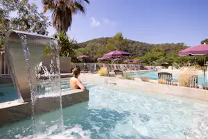 Camping-Pool mit Wasserdusche, Liegen und bunten Sonnenschirmen, im Hintergrund Bäume und ein bewaldeter Hügel.