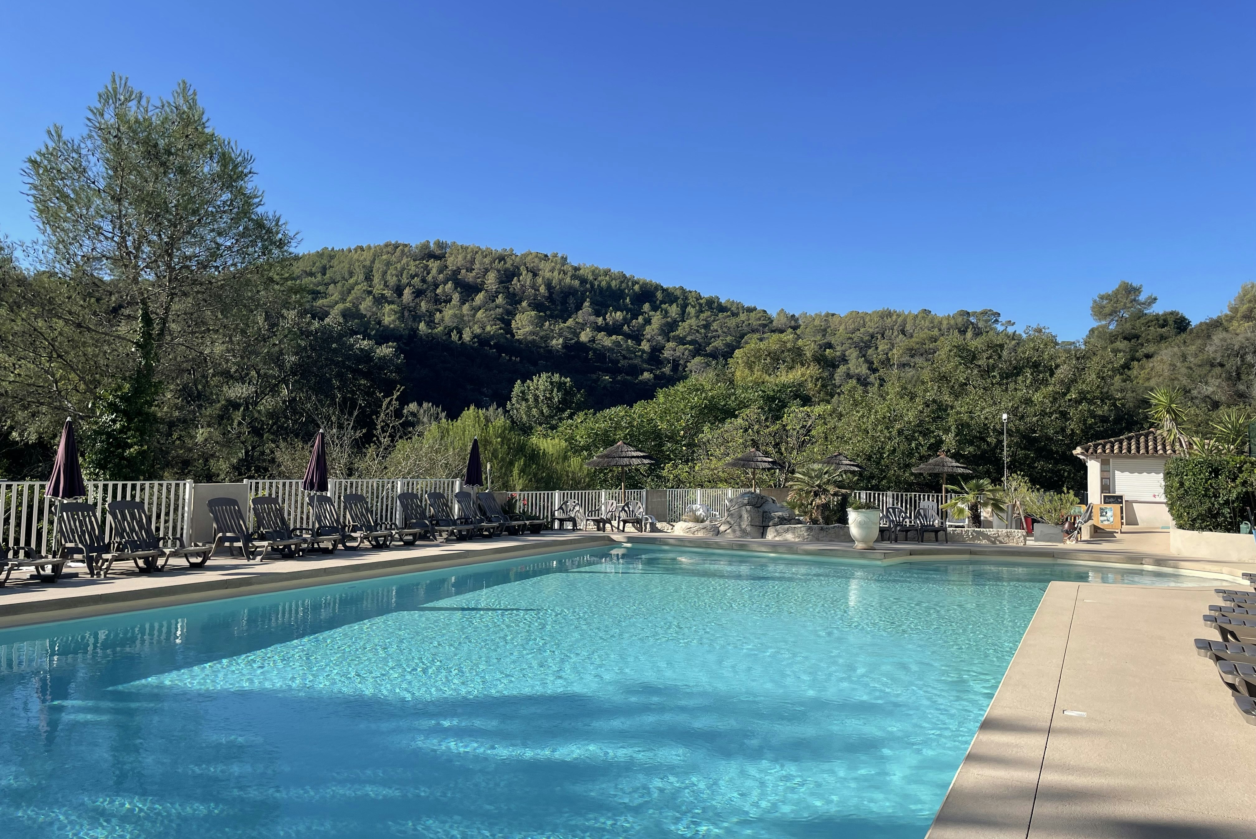 Schwimmbad mit türkisfarbenem Wasser, Liegen und Sonnenschirmen am Rand, dahinter Bäume und ein bewaldeter Hügel.