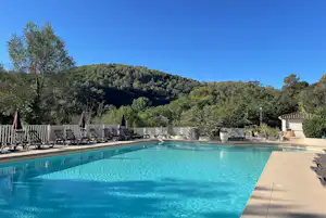 Schwimmbad mit türkisfarbenem Wasser, Liegen und Sonnenschirmen am Rand, dahinter Bäume und ein bewaldeter Hügel.