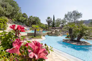 Blumen und Palmen vor einem Camping-Pool mit Liegen, Steinbecken und Wegen im Garten, umgeben von Bäumen und Hügeln.