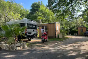 Wohnmobil auf einem Campingplatz-Stellplatz neben einem Holzhaus, im Hintergrund Bäume und ein Motorrad vor dem Weg.