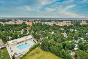 Luftaufnahme eines Campingplatzes mit Schwimmbecken, gepflegten Wegen und vielen Bäumen, im Hintergrund eine Küstenstadt.