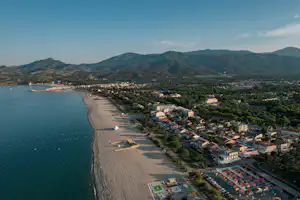 Luftaufnahme einer Küste mit Bucht, langem Strand und Campingbereich am Ufer, dahinter Häuser und bewaldete Berge.