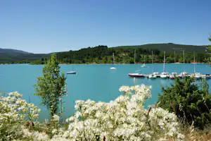 Türkisfarbenes Seeufer mit Liegeplatz: mehrere Segelboote im Hafen, umgeben von Bäumen und grünen Hügeln.
