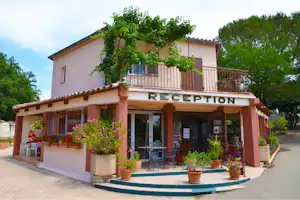 Camping-Rezeption mit Schild „RECEPTION“, Terrasse mit Geländer, Töpfen und Blumen vor dem Gebäude, umgeben von Bäumen.