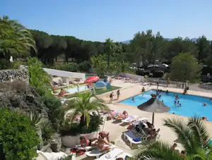 Schwimmbad mit Sonnenschirmen und Liegen auf dem Campingplatz, umgeben von Palmen, Bäumen und Wegen.