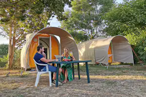 Zwei Campingzelte mit Vorraum auf einem Kiesplatz, davor ein Tisch mit zwei Personen und Stühlen, umgeben von Bäumen.