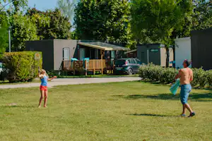 Mobilheim mit Holzterrasse und geparktem Auto am Campingplatz, umgeben von Bäumen und Rasenfläche.