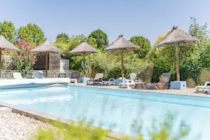 Schwimmbecken am Campingplatz mit Liegestühlen und Sonnenschirmen, im Hintergrund Hecken und mehrere Strohdächer.