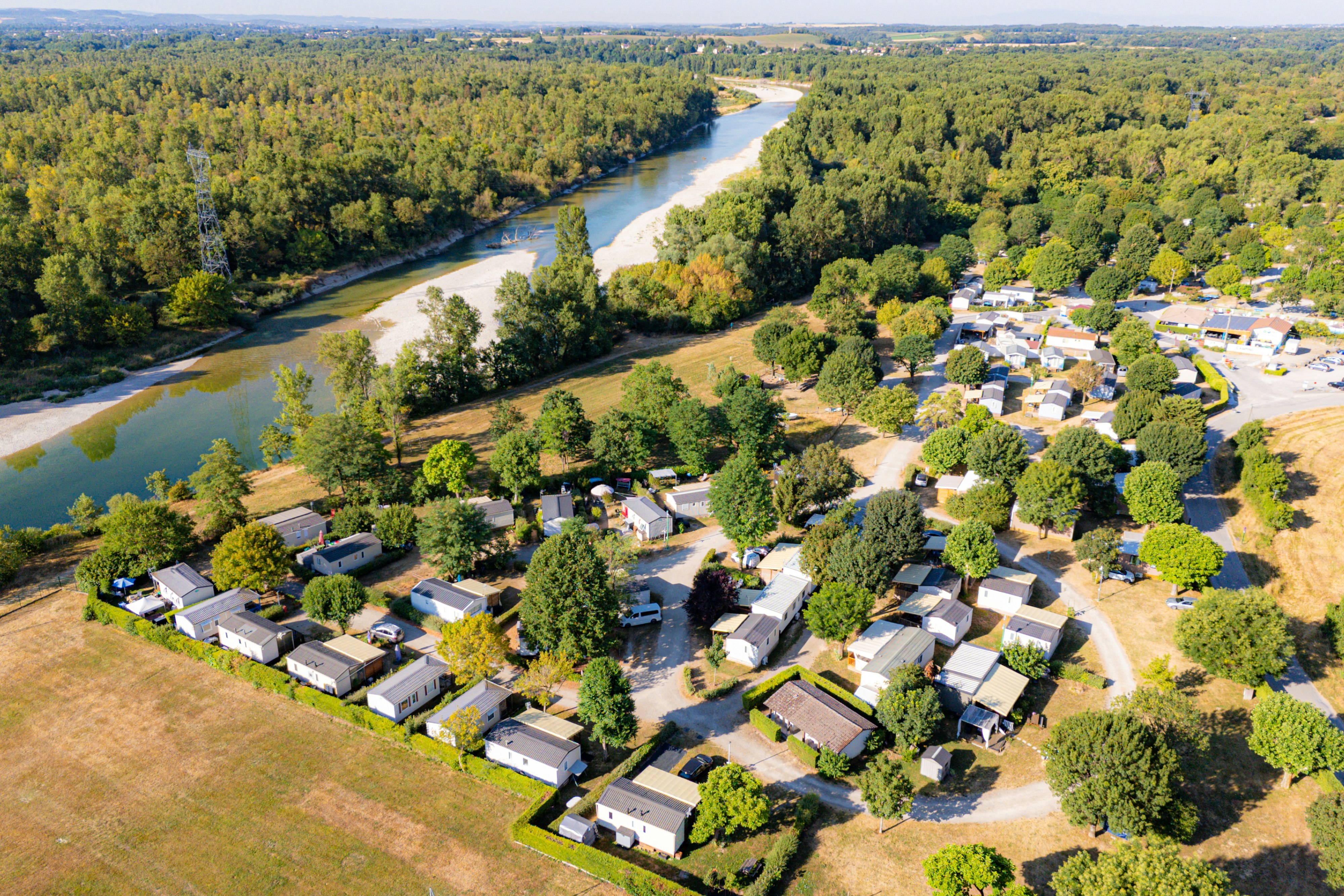 Luftaufnahme eines Campingplatzes mit Wohnwagen und Zelten, Wegen und Bäumen, am Fluss mit grünen Ufern.