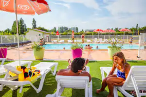 Poolbereich mit mehreren roten Sonnenschirmen, Liegen und zwei Personen auf dem Rasen vor dem Wasser.
