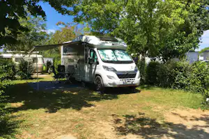 Wohnmobil auf einem Campingplatz-Stellplatz mit Vorzelt, Sonnenschirm und Sitzbank unter Bäumen.