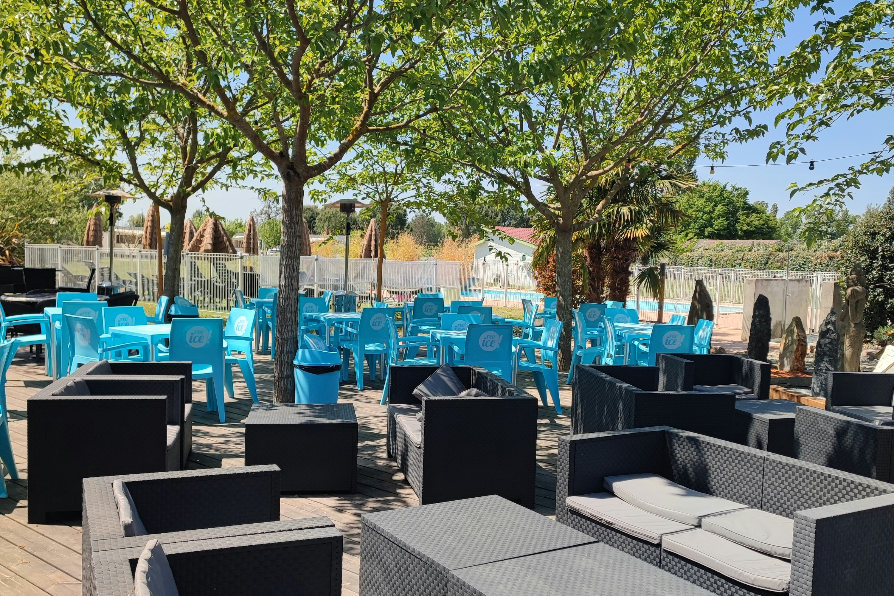 Terrasse mit modernen Lounge-Sesseln und Tischen, dahinter blaue Stühle und Bäume vor einem Zaun.