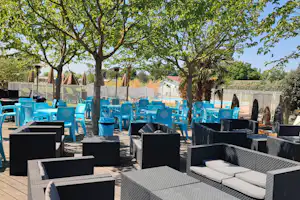 Terrasse mit modernen Lounge-Sesseln und Tischen, dahinter blaue Stühle und Bäume vor einem Zaun.