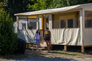 Zwei Personen vor einem Wohnwagen mit Vorzelt und großen Fenstern, am Stellplatz mit Holzpodest und Bäumen im Hintergrund.