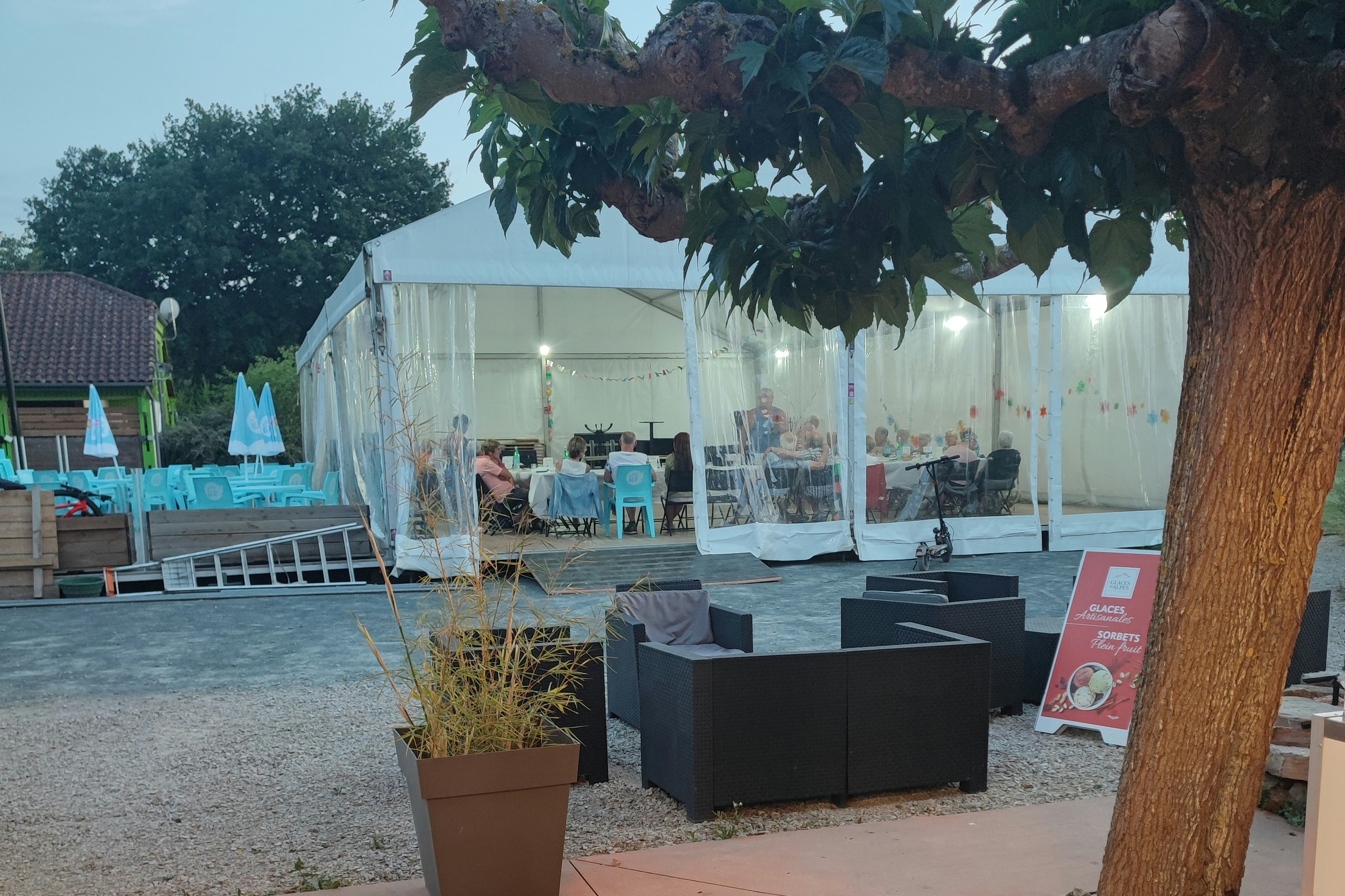 Terrasse mit Sitzbank und Tisch, daneben Pflanzkübel; im Hintergrund weiße Zelte mit beleuchteten Stühlen und Tischen.