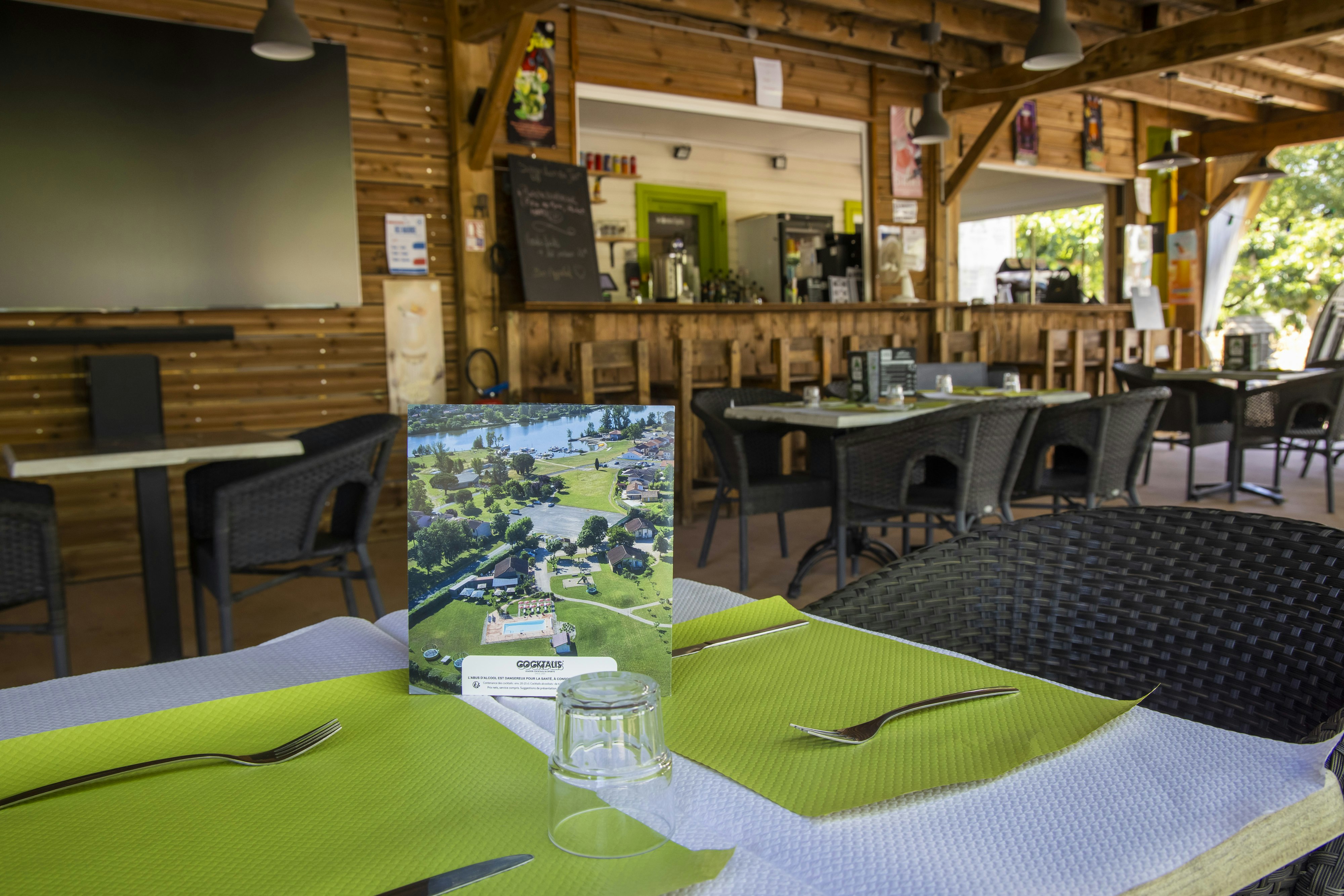 Grüner gedeckter Tisch im Camping-Restaurant, daneben Stühle und eine Karte, im Hintergrund Holztheke und Barbereich.