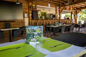 Grüner gedeckter Tisch im Camping-Restaurant, daneben Stühle und eine Karte, im Hintergrund Holztheke und Barbereich.