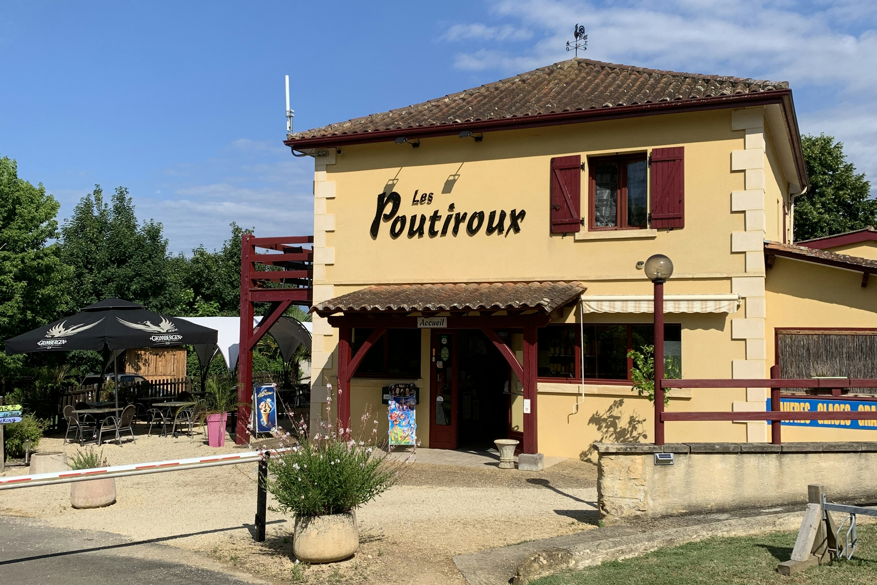 Gelbes Gebäude mit rotem Dach und Terrasse, Schild „Les Poutiroux“, davor Weg und Sitzbank im Campingbereich.
